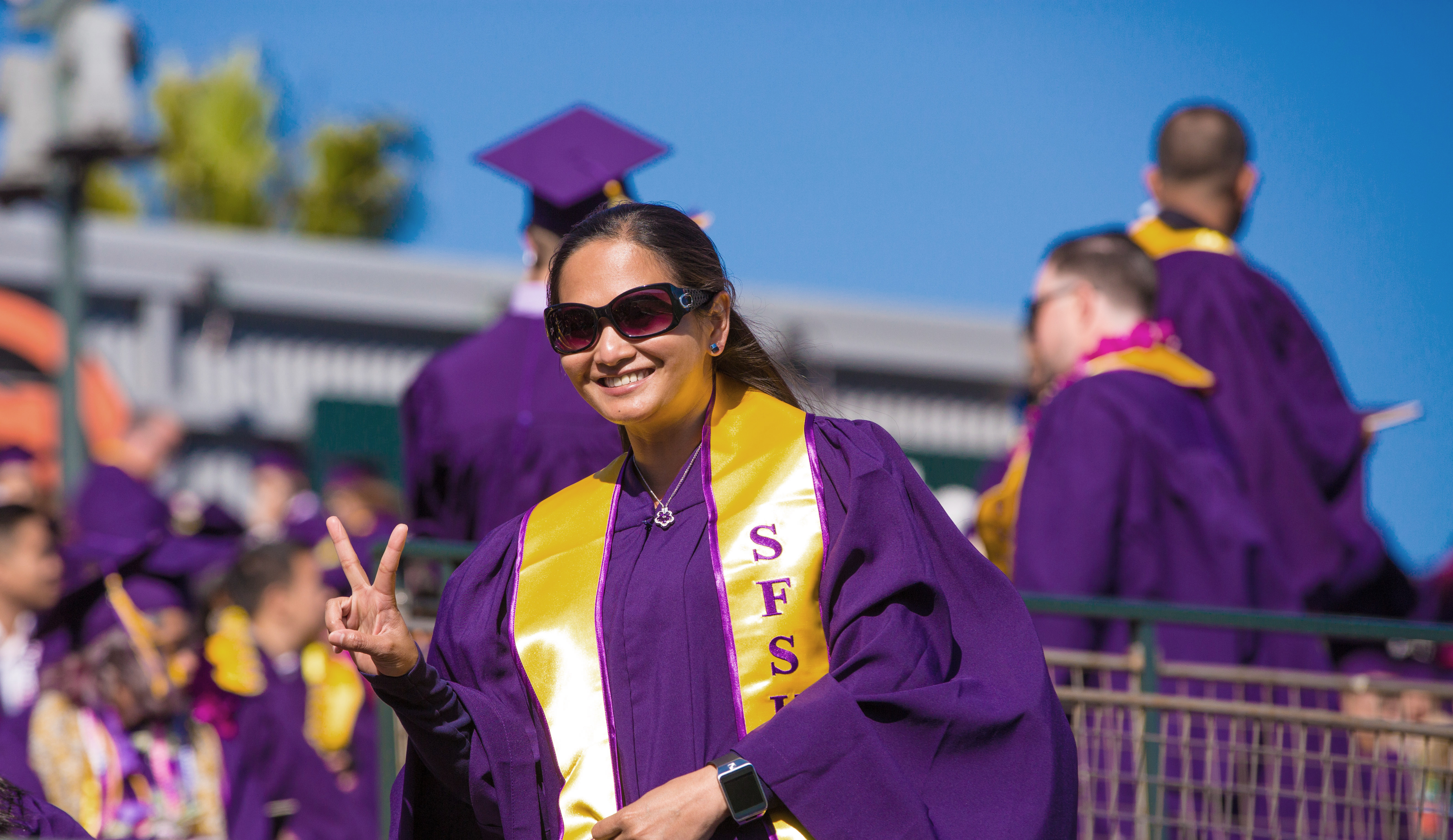 Female wearing sunglasses at graduation in purple gown showing a peace sign