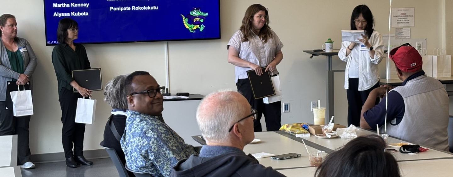 People in a classroom sitting around a table, while a person is being awarded