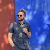 White man with beard and sunglasses standing in front of a red and blue wall